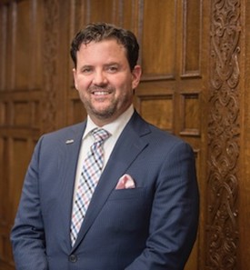 person standing against a wood panel background, wearing a dark grey suit and light grey tie with blue strips